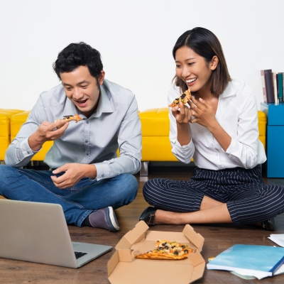 Cheerful Asian colleagues sitting on floor in office, eating pizza and watching funny video on laptop. Happy Asian couple or friends laughing at internet video and enjoying pizza together